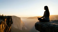 Woman meditating on mountain cliff edge at sunrise with scenic valley view below. Peaceful mindfulness practice in nature for wellness and spiritual growth.