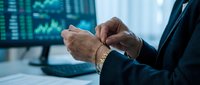 Mature male professional in dark business suit checking time on wristwatch at desk with financial data displayed on computer monitor in background.
