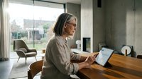 Mature Caucasian woman with gray hair and glasses using a tablet at a wooden table in a modern home office with garden view.
