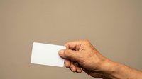 Senior male hand holding blank white business card against neutral beige background for corporate branding and professional networking purposes.