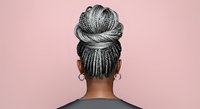 Young woman with dark skin tone wearing braided hairstyle in high bun with gold hoop earrings, photographed from behind against pink background for hair care and beauty content.