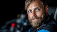 Confident mature Caucasian male with stylish beard and blue eyes wearing black jacket, looking away thoughtfully against dark background with soft lighting.