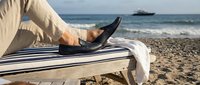 Man relaxing on beach chair wearing black shoes, feet up on striped lounger with ocean waves and cargo ship in background, vacation leisure lifestyle.