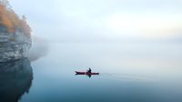 Lone kayaker paddling on misty lake at dawn, surrounded by limestone cliffs and autumn trees. Serene water reflects foggy atmosphere creating minimalist scene.