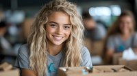 Young blonde woman with radiant smile wearing casual grey t-shirt in modern cafe setting, natural daylight illuminates authentic candid moment.