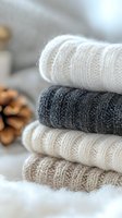 Stack of cozy knitted sweaters in neutral colors white, grey and beige on soft background with pine cone decoration, creating warm winter atmosphere.