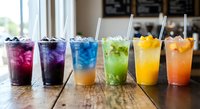 Colorful layered drinks with ice and straws arranged in rainbow order on wooden table in modern cafe setting for summer beverage menu design.