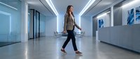 Woman in business casual attire walking through modern office lobby with glass partitions and contemporary lighting, suitable for corporate, workplace, and professional business concepts.
