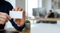 Man in dark blue shirt holding blank white business card in modern office workspace with computer and desk plant.