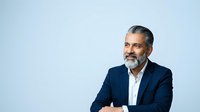 Middle-aged man with salt-and-pepper beard wearing navy blazer and white shirt against light blue background, suitable for corporate portraits and business headshots.