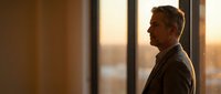 Mature businessman in suit looking out office window during golden hour sunset, professional executive contemplating future business decisions.