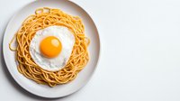 Cooked spaghetti pasta topped with sunny side up egg on round white ceramic plate against light background, overhead view of traditional breakfast meal.