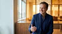 Confident middle-aged Caucasian businessman in navy suit smiling warmly in modern office with natural lighting and wooden interior elements.
