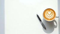 Coffee cup with latte art and pen on white paper background, overhead view workspace setup for business planning and creative writing projects.