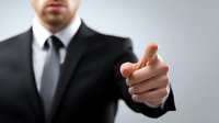 Businessman in formal suit pointing finger at viewer, conveying leadership and direct communication in professional setting against neutral background.