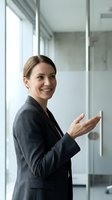 Professional businesswoman in gray suit presenting with open hand gesture in modern office environment with glass walls and natural lighting.