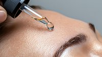 Closeup of serum drop falling from glass pipette onto face skin near eyebrow. Macro beauty shot showing moisturizing skincare product application.