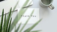 Thank you card on a white desk with green plant leaves, a mug, and laptop, creating a minimal and elegant workspace flat lay.