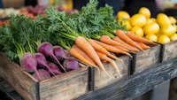 Fresh organic vegetables in rustic wooden crates at farmers market, including vibrant carrots with green tops and purple beets, with lemons in background.