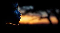 Silhouette profile of young African woman against dramatic sunset sky with glowing orange horizon and dark tree shapes creating atmospheric mood.