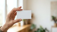 Male hand holding blank white business card against blurred interior background with natural window light, suitable for branding, corporate identity, and marketing mockup presentations.