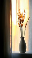 Silhouette of dried grass stems in ceramic vase on windowsill against soft golden sunlight through sheer curtain, creating peaceful minimalist home decor atmosphere.