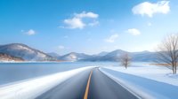 Scenic winter road along frozen lake with snow-covered mountains and bare trees, clear asphalt highway curves through white landscape under blue sky with clouds.