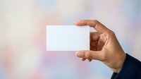 Hand holding blank white business card against soft blurred background, suitable for branding, corporate identity, and marketing mockup designs.