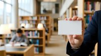 Businessman in dark suit holding blank white card in modern library or office setting with bookshelves and colleagues working in background for corporate branding.