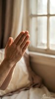 Hands clasped in prayer near a sunlit bedroom window, conveying faith, hope, and spiritual devotion in a warm, peaceful setting.