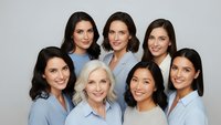 Diverse group of seven professional women in light blue uniforms smiling together in healthcare or corporate team portrait on neutral background.