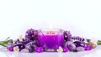 Purple candle surrounded by amethyst crystals and gemstones creating peaceful spa atmosphere for meditation and wellness rituals on white background.
