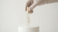Gloved hand placing elegant glass serum bottle with gold cap on white podium, minimalist beauty product presentation on light background.