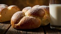 Fresh baked challah bread rolls with sesame seeds on rustic wooden table alongside glass of milk for breakfast or dinner meal preparation.