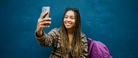 Young African American woman with dreadlocks taking selfie with smartphone against blue wall, wearing casual jacket with purple backpack.