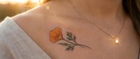 Closeup of orange flower tattoo on woman's chest with golden sunlight and delicate necklace, suitable for beauty and body art content.