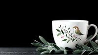 White ceramic cup with painted bird and floral design on black background with fresh sage leaves, moody minimalist composition for food and beverage styling.
