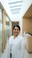 Young South Asian female doctor in white lab coat smiling confidently in a modern bright medical clinic hallway.