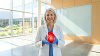 Senior female cardiologist with white hair, Caucasian ethnicity, wearing white medical coat and blue stethoscope, holding red heart model in modern bright clinic office with large windows and wooden accents, representing cardiovascular health, medical education, and cardiology practice.