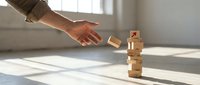 Young adult male hand reaching toward stacked wooden blocks in bright, minimalist interior space with natural window light, representing risk, strategy, and decision-making concepts.