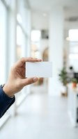 Hand holding blank white business card in modern office environment with blurred background, suitable for branding and corporate identity mockups.