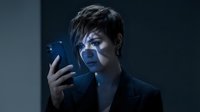 Middle-aged Asian woman in dark business attire holding smartphone with blue light illuminating her face against dark background for technology and communication concepts.