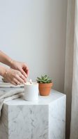 Woman hands lighting white candle next to small succulent plant on marble table in minimalist home interior for relaxation and wellness lifestyle.