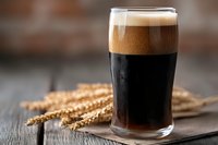 Glass of dark craft beer with thick foam head and wheat ears on rustic wooden table, selective focus on fresh cold stout or porter brew.