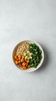 Healthy vegan grain bowl with roasted sweet potato, quinoa, kale, avocado, and seeds on a light gray background, top view.