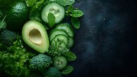 Fresh green vegetables including avocado, broccoli, cucumber slices and mint leaves arranged on dark background for healthy eating and nutrition concepts.
