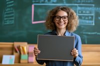 Young female teacher with curly hair and glasses holding blank chalkboard while standing in classroom with green chalkboard background. Cheerful expression for education concept.