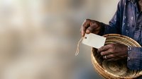 Adult male with dark skin tone wearing denim shirt holding empty wallet and straw hat, representing financial hardship, poverty, and economic struggle for editorial and awareness content.