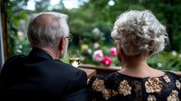 Senior Caucasian couple enjoying wine on patio, woman in floral dress and man in dark suit viewing garden with pink roses, romantic outdoor moment.
