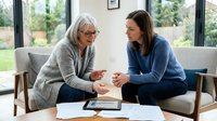 Senior woman with white hair and glasses wearing gray cardigan speaking with younger adult woman in blue shirt during counseling or mentoring session at home with tablet on table.